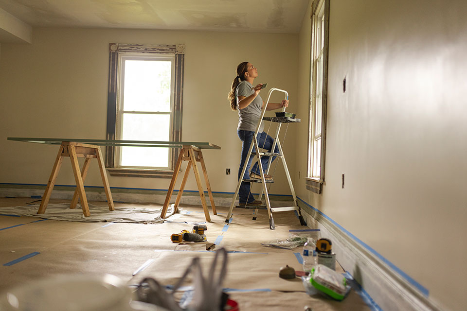 A woman steps up a ladder with a paint brush to paint the wall.