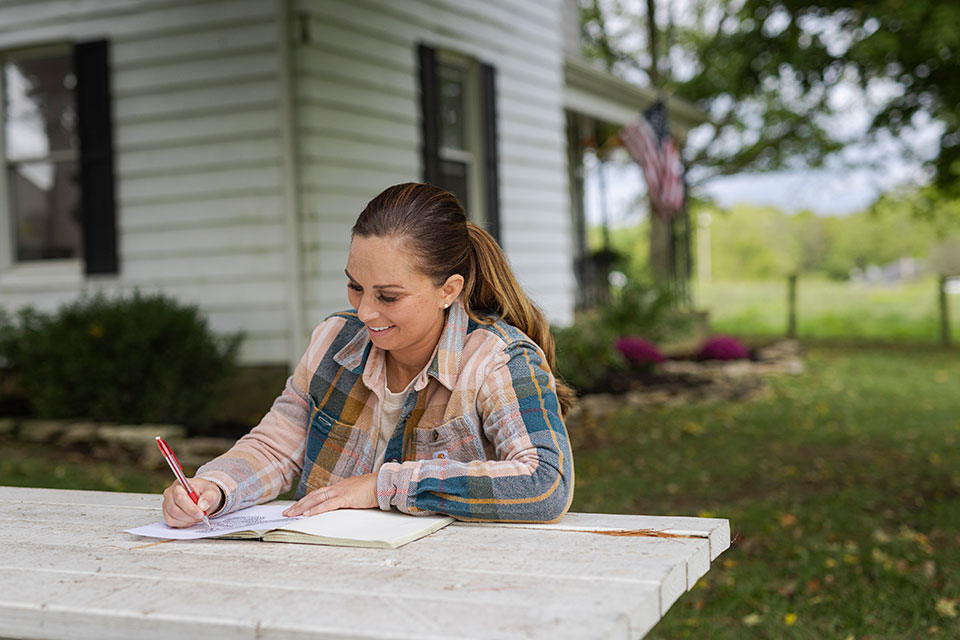 A woman writes in a notebook on a picnic bench outside a home.